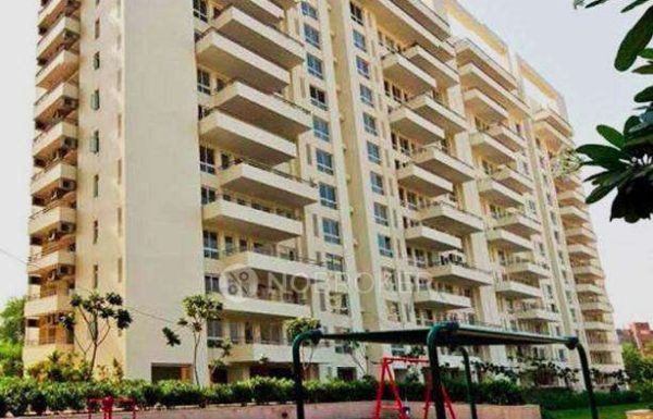 A tall cream-colored residential apartment building with multiple spacious balconies, large windows, landscaped greenery, and a children’s swing play area in the foreground, captured during daylight.