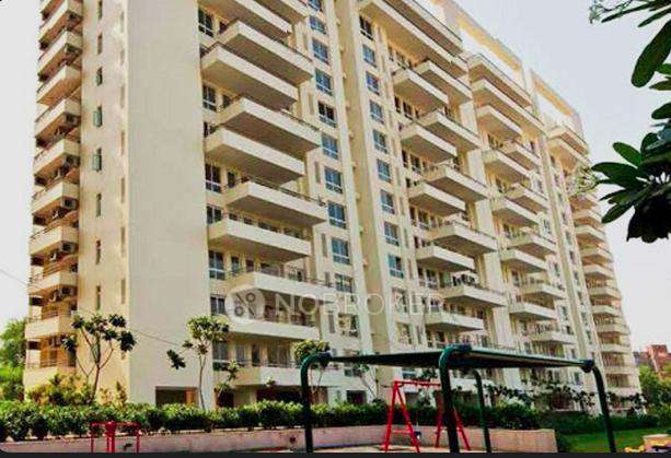 A tall cream-colored residential apartment building with multiple spacious balconies, large windows, landscaped greenery, and a children’s swing play area in the foreground, captured during daylight.